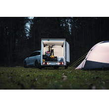 Humbaur Kofferanhänger mit geöffneten Türen, beladen mit einem Fahrrad und Campingausrüstung, neben einem Zelt auf einer Wiese im Wald.