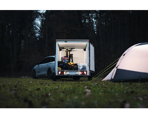 Humbaur Kofferanhänger mit geöffneten Türen, beladen mit einem Fahrrad und Campingausrüstung, neben einem Zelt auf einer Wiese im Wald.