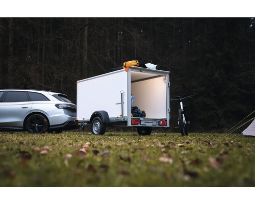 Weißer Humbaur Kofferanhänger an einem Auto auf einer Waldwiese, mit geöffneter Hecktür, Innenbeleuchtung und Campingausrüstung.
