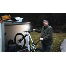 Ein Mann steht im Freien neben einem beleuchteten Kastenanhänger und hält ein weißes Mountainbike fest.