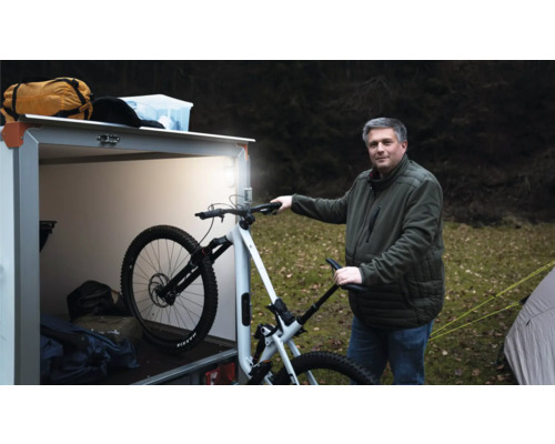 Ein Mann steht im Freien neben einem beleuchteten Kastenanhänger und hält ein weißes Mountainbike fest.