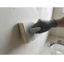 Hand mit Arbeitshandschuh nutzt ein Schwammbrett zum Glätten von Putz an einer Wand.