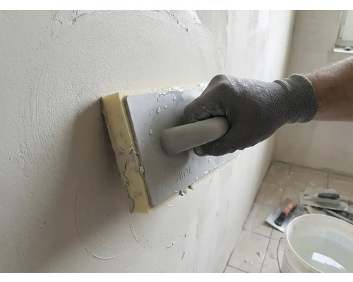 Hand mit Arbeitshandschuh nutzt ein Schwammbrett zum Glätten von Putz an einer Wand.