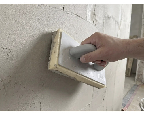 Hand führt ein Schwammbrett 28 mal 14 Zentimeter zum Glätten von frischem Putz über eine Wand.