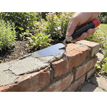 Hand mit Maurerlob Maurerkelle trägt Mörtel auf eine Ziegelmauer im Garten auf.