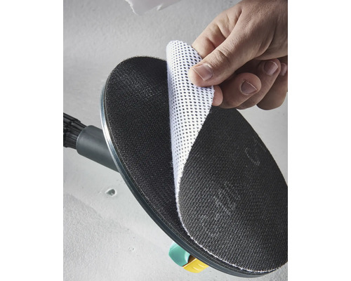 Eine Hand befestigt eine weiße Gitterschleifscheibe auf dem schwarzen runden Schleifteller eines Trockenbauschleifers.