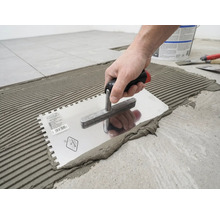 Eine Hand trägt mit einer gezahnten Glättkelle Fliesenkleber in einem Rillenmuster auf den Boden auf.