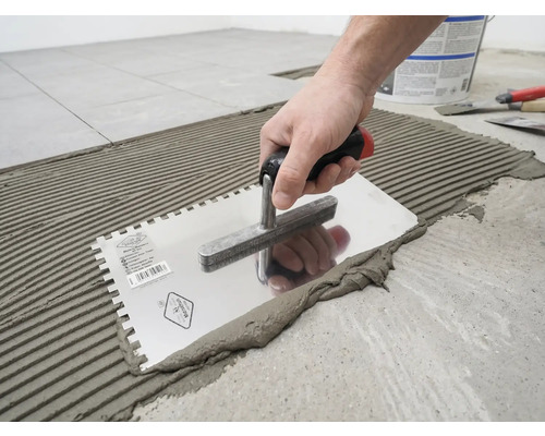 Eine Hand trägt mit einer gezahnten Glättkelle Fliesenkleber in einem Rillenmuster auf den Boden auf.