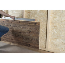Eine Person richtet ein Wandpaneel in Holzoptik mit einer Wasserwaage an einer Wand aus Grobspanplatten aus.