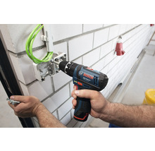 Bosch Professional Akku-Bohrschrauber befestigt eine Elektro-Verteilerdose mit grünen Kabeln an einer weißen Ziegelwand.