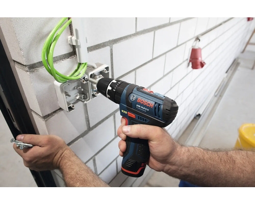 Bosch Professional Akku-Bohrschrauber befestigt eine Elektro-Verteilerdose mit grünen Kabeln an einer weißen Ziegelwand.