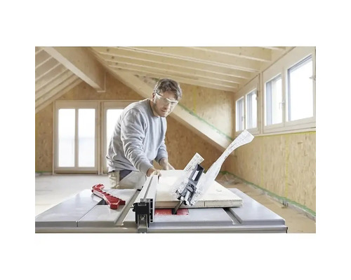 Ein Mann mit Schutzbrille schneidet ein Holzbrett mit einer Tischkreissäge in einem hellen Raum mit Holzbalken und OSB-Wänden.