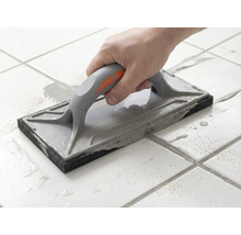 Eine Hand führt ein Fugenbrett über weiße Bodenfliesen, um graue Fugenmasse zu verteilen.