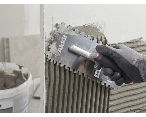 Eine Hand mit Handschuh trägt mit einer Akkit Glättkelle Fliesenkleber gleichmäßig auf eine Wand auf.