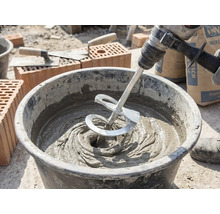 Ein Rührwerk an einer Bohrmaschine mischt grauen Mörtel in einem Baueimer auf einer Baustelle mit Ziegeln und Maurerkellen.