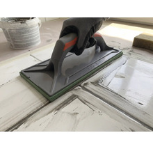 Hand mit Schutzhandschuh nutzt ein Fugenbrett zum Verfugen von Fliesen.