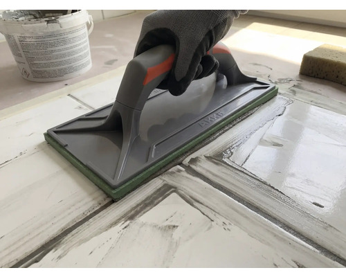 Hand mit Schutzhandschuh nutzt ein Fugenbrett zum Verfugen von Fliesen.