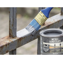 Ein Pinsel trägt grauen Metallschutzlack auf einen rostigen Metallzaun auf. Daneben steht eine Dose 3 in 1 Metallschutzlack Mattlack.