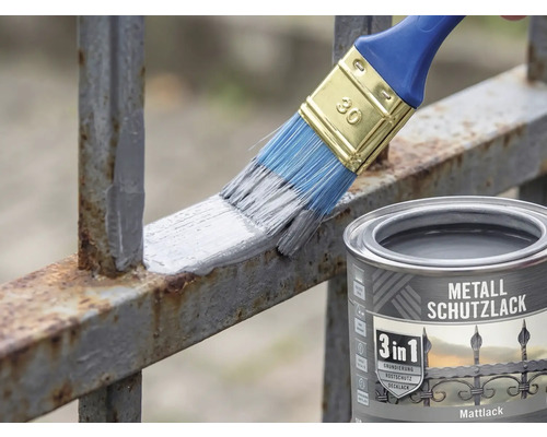 Ein Pinsel trägt grauen Metallschutzlack auf einen rostigen Metallzaun auf. Daneben steht eine Dose 3 in 1 Metallschutzlack Mattlack.