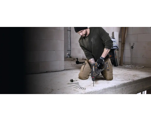 Handwerker in Arbeitskleidung bohrt mit einem Worx Akkumulator-Bohrhammer Löcher in einen Betonboden auf einer hellen Baustelle.