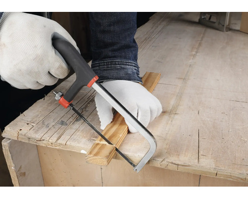 Person mit weißen Schutzhandschuhen schneidet eine Holzleiste mit einer kleinen Handsäge auf einer hölzernen Werkbank.