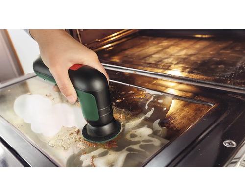 Eine Hand reinigt die Glasscheibe einer Backofentür mit einer elektrischen Akku-Reinigungsbürste und Reinigungsschaum.