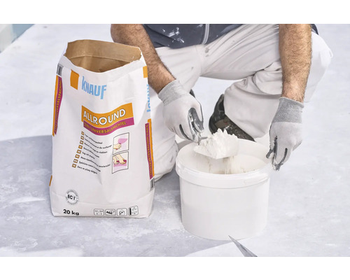 Handwerker entnimmt Spachtelmasse aus Kübel neben einem 20 Kilogramm Sack Knauf Allround 2 in 1 Universalspachtel.