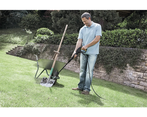 Ein Mann reinigt eine verschmutzte Schaufel in einer Scheibtruhe mit einem Hochdruckreiniger auf einer Rasenfläche im Garten.