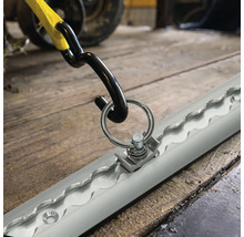 Zurrschiene mit Endbeschlag aus Metall und Haken eines Spanngurts zur Ladungssicherung auf einem Holzboden.