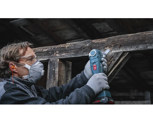Handwerker mit Schutzausrüstung verwendet ein Akku Multitool zum Sägen an einem alten Holzbalken.