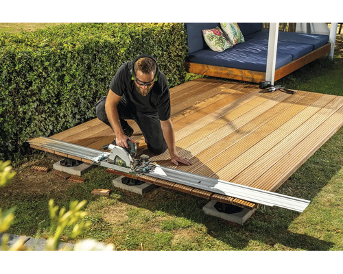Ein Handwerker schneidet Holz-Terrassendielen mit einer Handkreissäge und Führungsschiene im Garten.