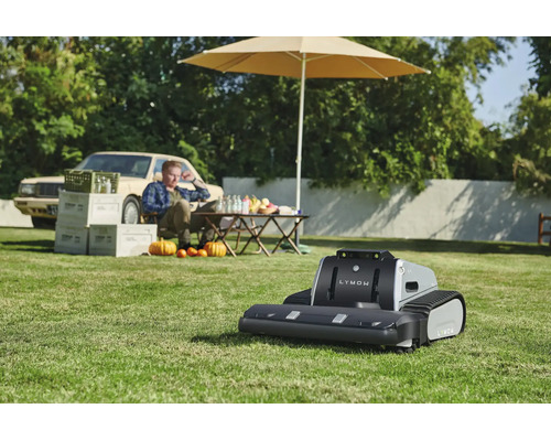 Mähroboter Lymow mit Kettenantrieb auf einer Rasenfläche in einem sonnigen Garten mit Tisch, Sonnenschirm und einer Person im Hintergrund.