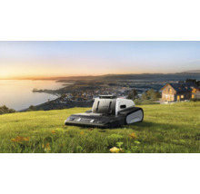 Lymow Mähroboter mit Raupenantrieb auf einer Rasenfläche mit Ausblick auf eine Küstenstadt bei Sonnenuntergang.