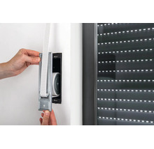 Hände montieren einen metallenen Gurtwickler für Rollladen in eine Wandöffnung neben einem Fenster.