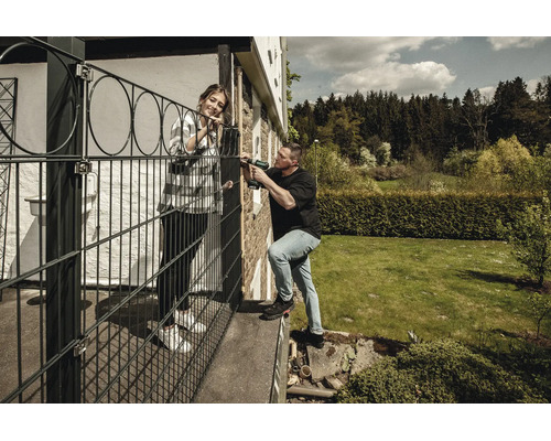 Ein Mann montiert einen schwarzen Schmuckzaun auf einer Terrasse mit einem Akkuschrauber, während eine Frau im sonnigen Garten zuschaut.