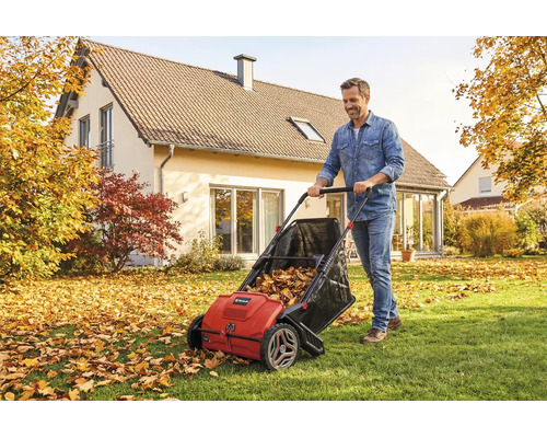 Ein Mann schiebt einen roten Laub- und Grassammler über eine Rasenfläche mit herbstlichem Laub vor einem Einfamilienhaus in einer hellen Umgebung.