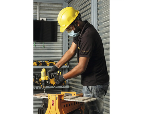 Handwerker mit Schutzhelm nutzt DeWALT Akku-Bohrschrauber an einem Sägebock in einer hellen Werkstatt mit Metallwänden.