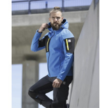 Ein bärtiger Mann trägt eine blau-schwarze Softshelljacke mit Kapuze, gelben Reißverschlüssen und Mascot Logo sowie eine schwarze Arbeitshose im Freien.
