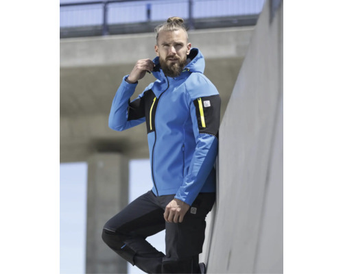 Ein bärtiger Mann trägt eine blau-schwarze Softshelljacke mit Kapuze, gelben Reißverschlüssen und Mascot Logo sowie eine schwarze Arbeitshose im Freien.