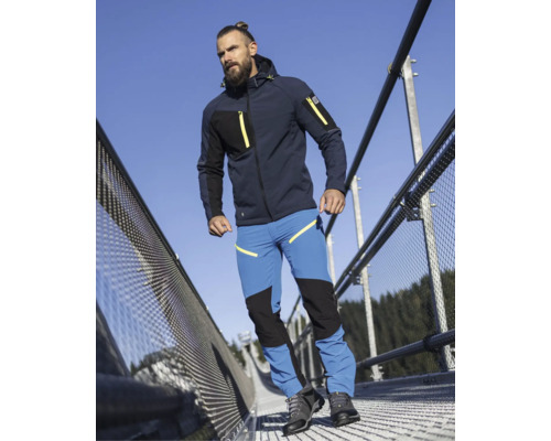 Mann in dunkelblauer Softshelljacke mit Kapuze und blauer Arbeitshose mit schwarzen Verstärkungen auf einer Metallbrücke.