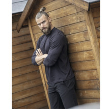 Ein Mann in dunkler Arbeitskleidung lehnt mit verschränkten Armen an der Holzwand eines Gartenhauses.