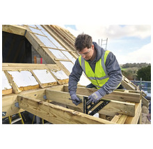 Handwerker markiert mit einem Sparrenwinkel und Bleistift einen Holzbalken auf einer hellen Dachbaustelle mit Holz- und Isoliermaterialien.