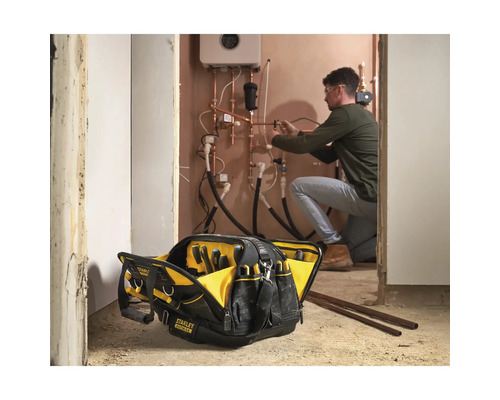Offene Stanley Fatmax Werkzeugtasche im Vordergrund, Handwerker arbeitet im Hintergrund an Heizungsrohren.