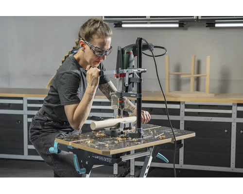 Person nutzt Bohrständer mit Bohrmaschine an einer Werkbank in einer hellen Werkstatt, trägt Schutzbrille und Gehörschutz.