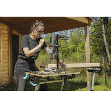 Eine Frau bohrt mit einem Bohrständer und einer Bohrmaschine in einen Holzbalken auf einer mobilen Werkbank im Garten.