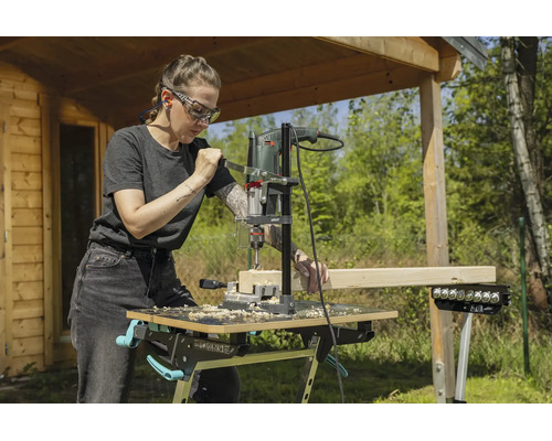 Eine Frau bohrt mit einem Bohrständer und einer Bohrmaschine in einen Holzbalken auf einer mobilen Werkbank im Garten.