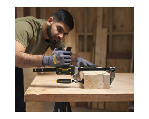 Stanley Fatmax Einhandzwinge fixiert Holzbalken auf einer Werkbank in einer hellen Werkstatt. Ein Mann mit Handschuhen bedient das Werkzeug.