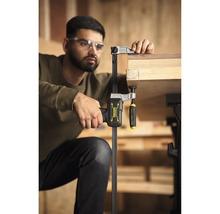 Handwerker mit Schutzbrille fixiert ein Holzbrett mit einer Stanley FatMax Einhandzwinge an einer Werkbank in einer hellen Werkstatt.
