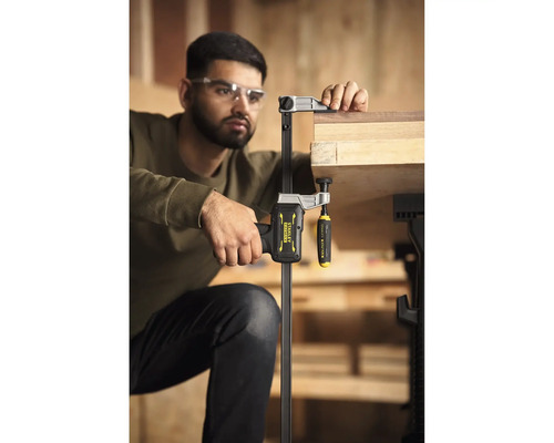 Handwerker mit Schutzbrille fixiert ein Holzbrett mit einer Stanley FatMax Einhandzwinge an einer Werkbank in einer hellen Werkstatt.