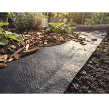 Schwarzes Unkrautvlies in einem Gartenbeet, teilweise mit Rindenmulch bedeckt, neben Erde und kleinen Pflanzen.